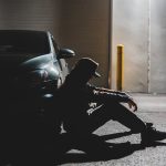 anonymous sad man sitting on ground near parked contemporary car in evening